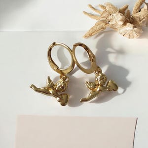 May include: A pair of gold-colored hoop earrings. Each earring features a small, detailed cherub pendant. The cherubs are depicted with wings and are holding a heart-shaped object. The earrings are displayed on a white surface with dried floral accents.
