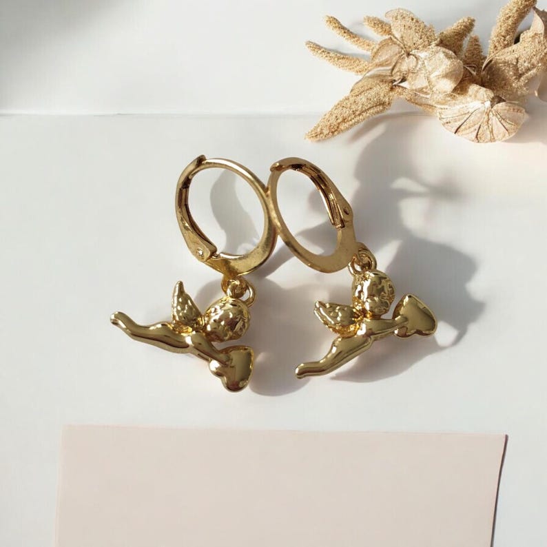 May include: A pair of gold-colored hoop earrings. Each earring features a small, detailed cherub pendant. The cherubs are depicted with wings and are holding a heart-shaped object. The earrings are displayed on a white surface with dried floral accents.