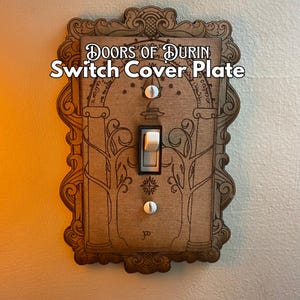 Peut inclure: Plaque de recouvrement d'interrupteur en bois avec le texte "Doors of Durin Switch Cover Plate". La plaque présente une bordure ornée et un motif détaillé d'arbres et d'arches. L'interrupteur est blanc et la plaque est fixée au mur.