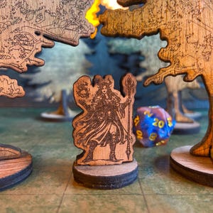 Puede incluir: Piezas de juego de mesa de madera, que incluyen una figura detallada de un personaje con un bastón, ambientada en una escena de bosque. Las piezas están cortadas con láser de madera, con diseños intrincados. También se ve un dado de veinte caras azul y morado.