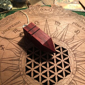 Könnte beinhalten: Ein Holzpendel mit roter Oberfläche und einer daran befestigten Schnur. Das Pendel liegt auf einem Holzbrett mit einem kreisförmigen Design und den aufgedruckten Wörtern "Oui" und "Plus".