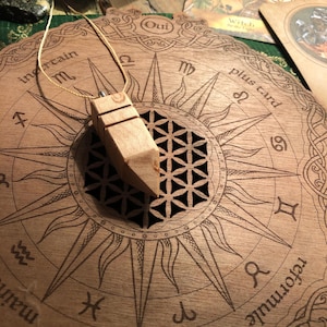 Könnte beinhalten: Ein Holzpendel mit einem geschnitzten Design hängt an einer Schnur über einem runden Holzbrett mit einem Lebensblume-Design und Tierkreiszeichen. Das Brett hat die Worte "Oui" und "plus card" darauf geschrieben.