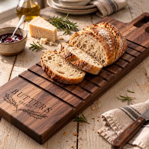 May include: A rectangular dark brown wooden bread board featuring a handle with a hole and horizontal grooves across the surface. The board is engraved with the text BUTTERS BAKERY and a wheat stalk illustration, holding several slices of crusty bread.
