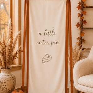 May include: A vertical cream-colored fabric banner suspended from a wooden frame with long, dark orange satin ribbons tied in bows at the top corners. The banner features the text "a little cutie pie" in dark brown script above a line-art pie slice.
