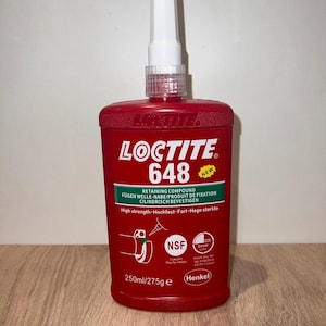 Puede incluir: Una botella roja de compuesto de retención Loctite 648 con una tapa blanca y una etiqueta verde. La botella está etiquetada como "Nuevo" y tiene el texto "High strength-Hochfest-Fort-Hoge sterkte". La botella es de 250 ml/275 g.