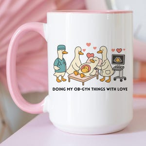 May include: White ceramic mug with a pink handle and rim. The mug features a cartoon illustration of ducks in medical attire performing an ultrasound. The text "DOING MY OB-GYN THINGS WITH LOVE" is printed below the illustration.