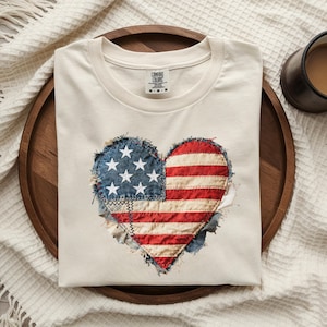 May include: Cream-colored t-shirt with a heart-shaped American flag design. The flag is made of denim and red and white striped fabric, with white stars on a blue background. The shirt is folded on a wooden tray.