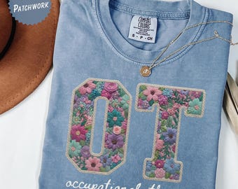Op de afbeelding: Een lichtblauw Comfort Colors T-shirt met de letters "OT" geborduurd met kleurrijke bloemen. De woorden "occupational therapy" staan eronder in het wit. Een gouden ketting met een hanger ligt over het shirt.
