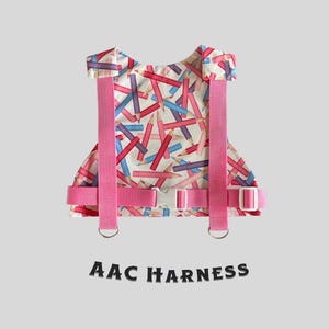 May include: Pink adjustable harness with a white background and a pattern of colourful pencils. The harness has two D-rings and a buckle. The text "AAC HARNESS" is printed below the harness.