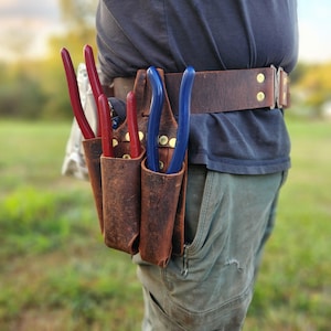 May include: A brown leather tool belt with three pockets, holding red and blue handled tools. The belt is worn around the waist and has brass rivets. The background is a blurred outdoor scene.