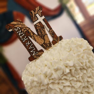 Op de afbeelding: Een wit glazuur taart met twee houten letter "M" taarttoppers. Eén topper heeft de naam "MARKINS" erop gedrukt. De andere "M" heeft een wit kruisontwerp. De toppers hebben een bruin, beige en wit kleurenschema.