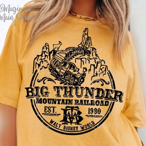Peut inclure: Un t-shirt jaune avec un motif noir d'un train montant une montagne avec le texte "Big Thunder Mountain Railroad Est. 1980 Walt Disney World"