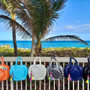 May include: Six colorful beach totes with a tennis racket design. The totes are lined up on a white railing with a beach and ocean in the background.