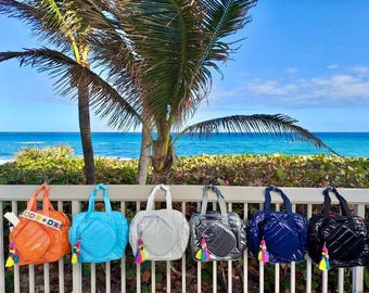 May include: Six colorful beach totes with a tennis racket design. The totes are lined up on a white railing with a beach and ocean in the background.
