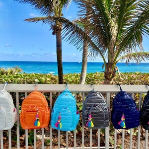 May include: Six quilted nylon backpacks in different colors hanging on a white railing. The backpacks have a zipper closure and a small pocket on the front. Each backpack has a colorful tassel hanging from the zipper pull.