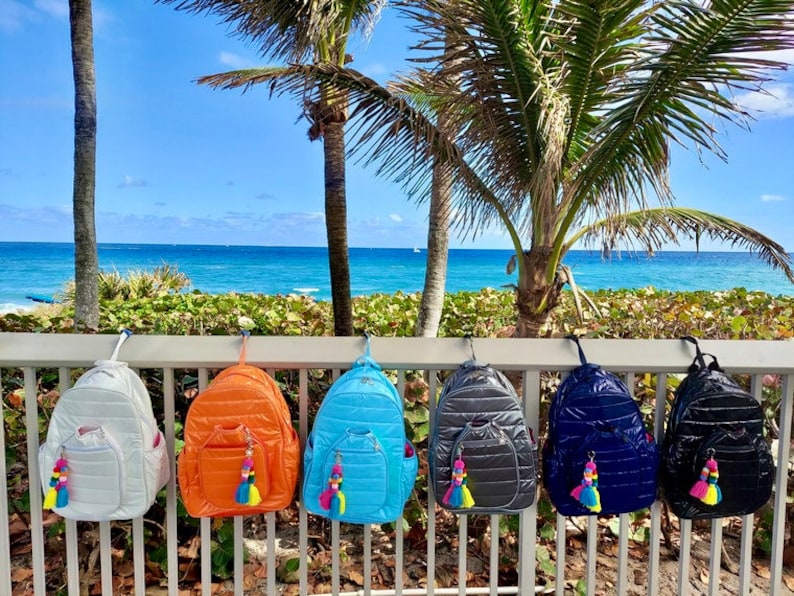 May include: Six quilted nylon backpacks in different colors hanging on a white railing. The backpacks have a zipper closure and a small pocket on the front. Each backpack has a colorful tassel hanging from the zipper pull.