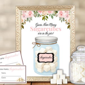 Op de afbeelding: Een ingelijst bord met bloemaccenten en de tekst "Raad hoeveel suikerklontjes er in de pot zitten!". Inclusief twee kleine kaarten om te raden en een glazen pot gevuld met suikerklontjes. Een decoratief element voor een feest.