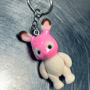 May include: A pink bunny keychain with a silver chain and ring. The bunny has a pink head and ears with brown tips, white eyes, and a beige body. The keychain is made of a soft, plush material.