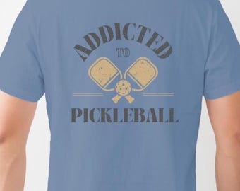 Może przedstawiać: Niebieska koszulka polo z napisem "Addicted to Pickleball" w kolorze czarnym i grafiką dwóch rakiet do pickleballa w kolorze beżowym.
