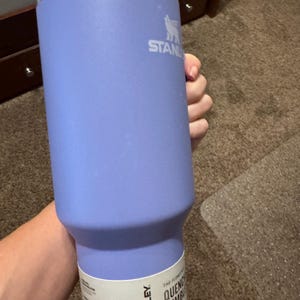 May include: A light purple Stanley Quencher tumbler with a 1.18 litre capacity. The stainless steel drinkware features a white paper label near the base with the Stanley logo and text, a silver metallic rim, and a white lid at the top of the vessel.