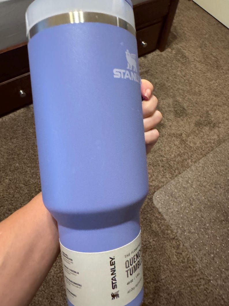 May include: A light purple Stanley Quencher tumbler with a 1.18 litre capacity. The stainless steel drinkware features a white paper label near the base with the Stanley logo and text, a silver metallic rim, and a white lid at the top of the vessel.