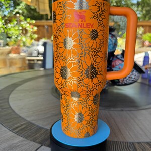 May include: An orange Stanley tumbler featuring a repeating sunflower pattern engraved in silver. The cup includes a matching orange handle, a white lid with an orange straw opening, and a silver rim. The brand name Stanley is printed in red.