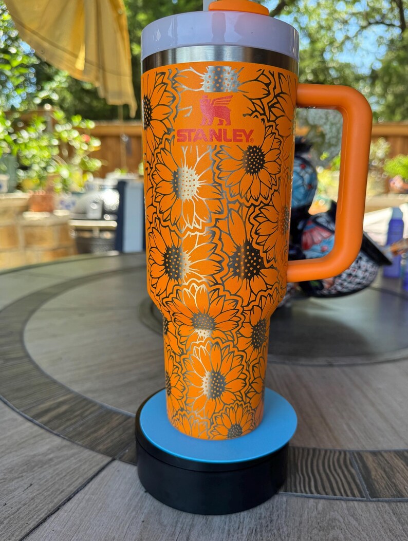 May include: An orange Stanley tumbler featuring a repeating sunflower pattern engraved in silver. The cup includes a matching orange handle, a white lid with an orange straw opening, and a silver rim. The brand name Stanley is printed in red.
