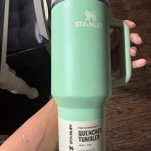May include: A light green Stanley Quencher Tumbler with a handle and a light purple lid. The tumbler features a white paper label with the text THE FLOWSTATE QUENCHER TUMBLER 40 OZ | 1.18 L All Day Hydration and the Stanley brand logo.
