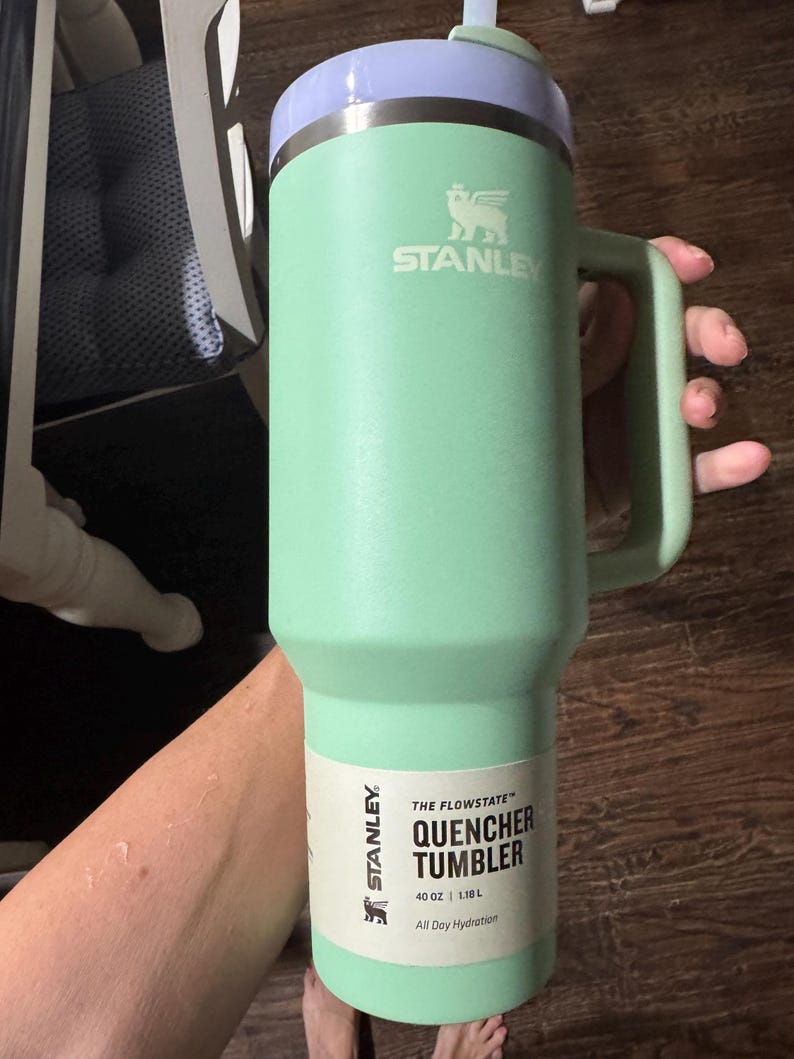 May include: A light green Stanley Quencher Tumbler with a handle and a light purple lid. The tumbler features a white paper label with the text THE FLOWSTATE QUENCHER TUMBLER 40 OZ | 1.18 L All Day Hydration and the Stanley brand logo.