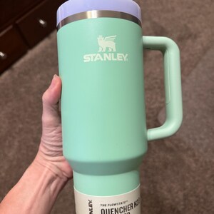 May include: A mint green Stanley Quencher H2.0 tumbler with a 1.18 L capacity. The cup features a light purple lid, a matching mint green handle, and a white straw. A product label at the base displays the text Stanley The Flowstate Quencher H2.0.