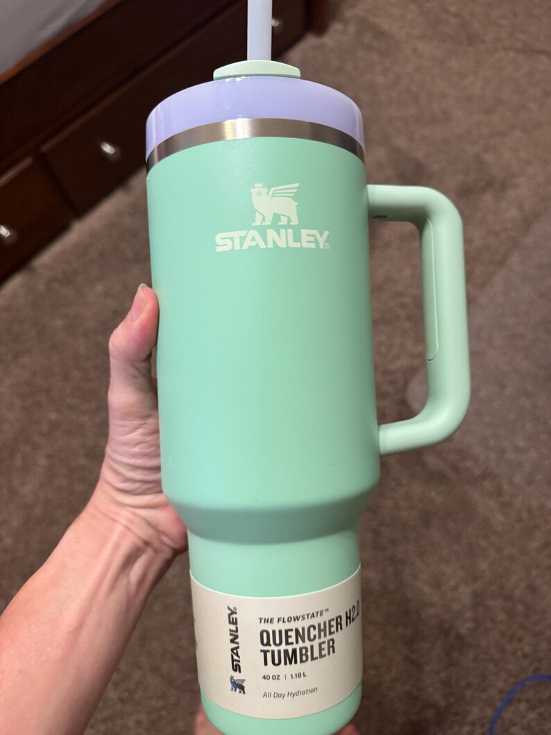 May include: A mint green Stanley Quencher H2.0 tumbler with a 1.18 L capacity. The cup features a light purple lid, a matching mint green handle, and a white straw. A product label at the base displays the text Stanley The Flowstate Quencher H2.0.