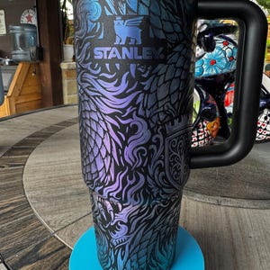 May include: A black Stanley tumbler featuring an iridescent purple and blue dragon-themed graphic print. The cup includes a large black side handle, a black lid, and the Stanley logo with a bear icon printed in the center of the upper section.