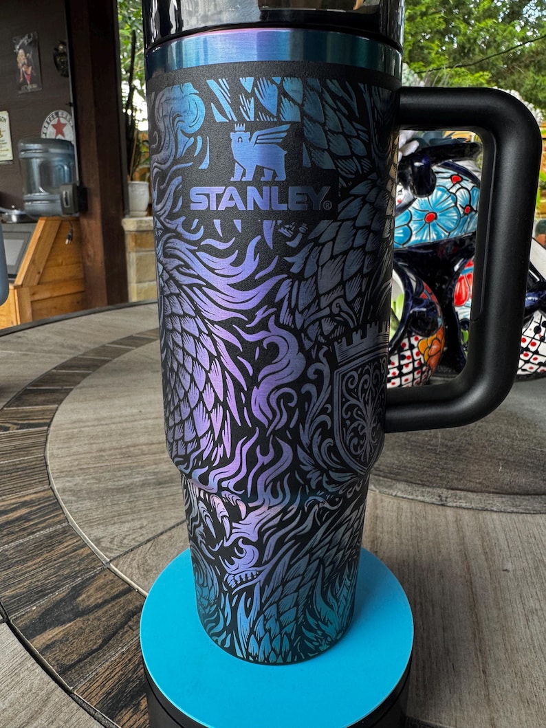 May include: A black Stanley tumbler featuring an iridescent purple and blue dragon-themed graphic print. The cup includes a large black side handle, a black lid, and the Stanley logo with a bear icon printed in the center of the upper section.