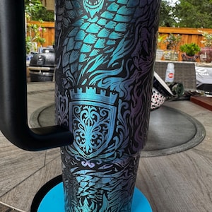 May include: A tall, cylindrical stainless steel tumbler with a matte black finish featuring a vibrant, iridescent teal and purple dragon scale pattern. The design includes a detailed crest emblem and a sturdy black handle attached to the side.
