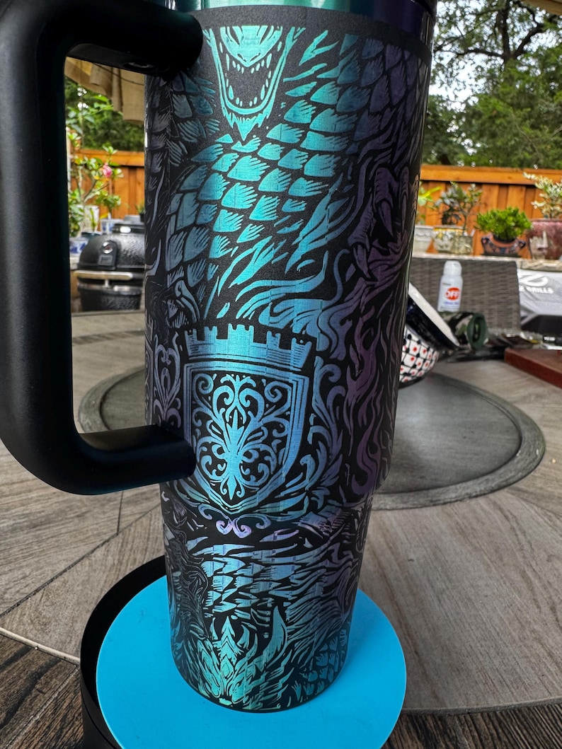 May include: A tall, cylindrical stainless steel tumbler with a matte black finish featuring a vibrant, iridescent teal and purple dragon scale pattern. The design includes a detailed crest emblem and a sturdy black handle attached to the side.