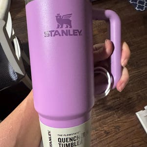 May include: A lavender-coloured Stanley Quencher tumbler with a handle and a clear lid. The cup features the Stanley logo and a paper label that reads The Flowstate Quencher Tumbler 30 oz .88 L All Day Hydration. The tumbler has a metallic rim.