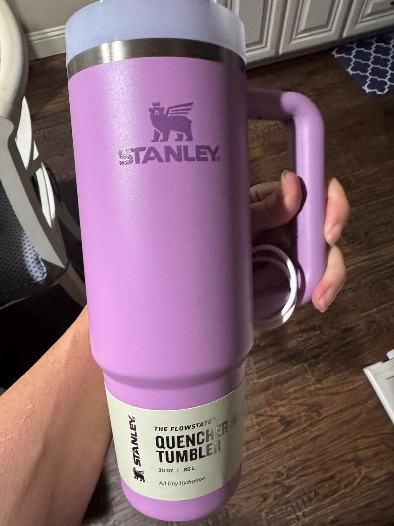 May include: A lavender-coloured Stanley Quencher tumbler with a handle and a clear lid. The cup features the Stanley logo and a paper label that reads The Flowstate Quencher Tumbler 30 oz .88 L All Day Hydration. The tumbler has a metallic rim.