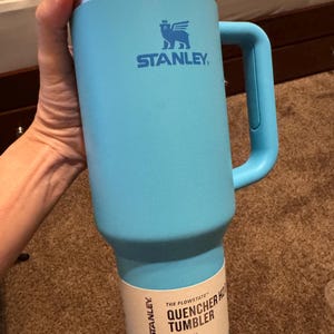 May include: A light blue Stanley Quencher H2.0 FlowState tumbler with a handle and a clear lid. The cup features the Stanley logo and a paper label that reads The Flowstate Quencher H2.0 Tumbler 40 oz 1.18 L All Day Hydration.