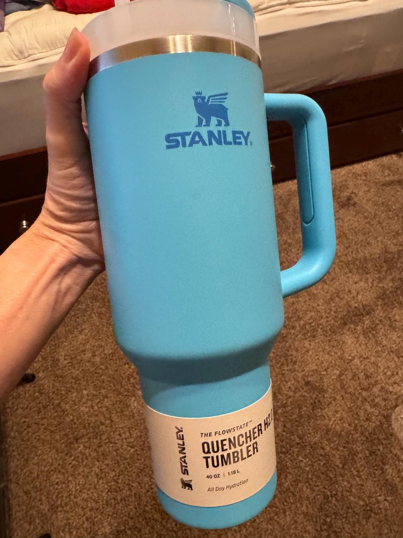 May include: A light blue Stanley Quencher H2.0 FlowState tumbler with a handle and a clear lid. The cup features the Stanley logo and a paper label that reads The Flowstate Quencher H2.0 Tumbler 40 oz 1.18 L All Day Hydration.