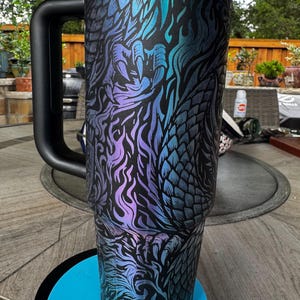 May include: A tall, cylindrical stainless steel tumbler with a black handle and a black lid. The exterior features a matte black finish with an intricate, iridescent dragon scale and flame pattern in shades of blue, purple, and teal.