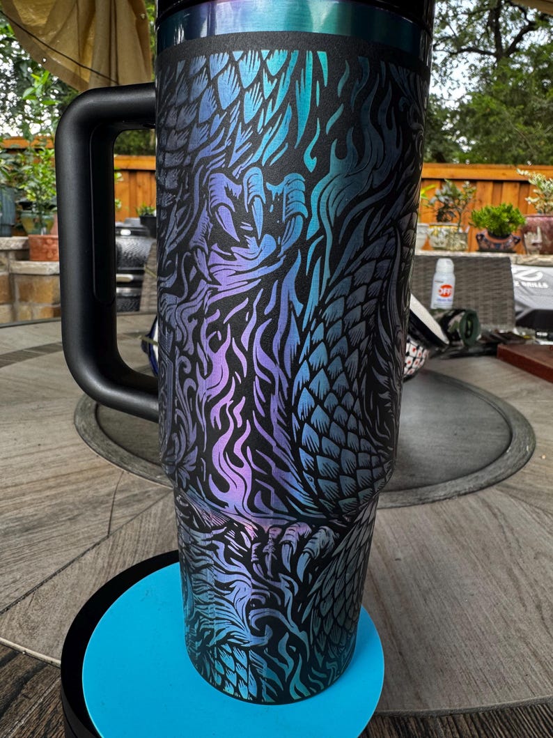 May include: A tall, cylindrical stainless steel tumbler with a black handle and a black lid. The exterior features a matte black finish with an intricate, iridescent dragon scale and flame pattern in shades of blue, purple, and teal.