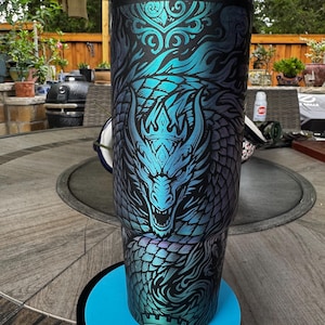 May include: A tall, cylindrical stainless steel tumbler featuring a black base color with a vibrant, iridescent teal and blue dragon graphic. The design includes intricate scale patterns, swirling smoke-like motifs, and a crown detail. It has a black lid.