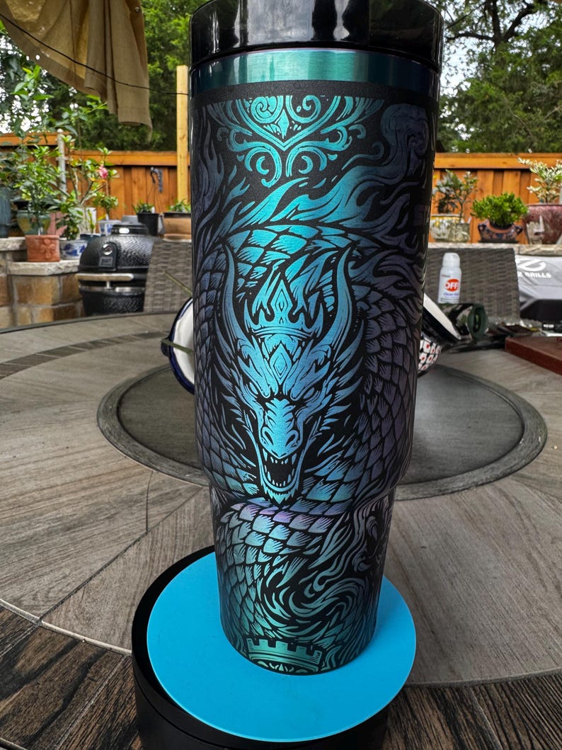 May include: A tall, cylindrical stainless steel tumbler featuring a black base color with a vibrant, iridescent teal and blue dragon graphic. The design includes intricate scale patterns, swirling smoke-like motifs, and a crown detail. It has a black lid.