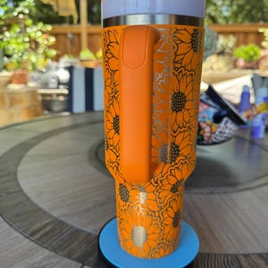 May include: A bright orange tumbler featuring a repeating sunflower pattern in a metallic silver finish. The cup includes a sturdy orange side handle, a white lid, and a clear straw. It sits upright on a circular blue and black base.