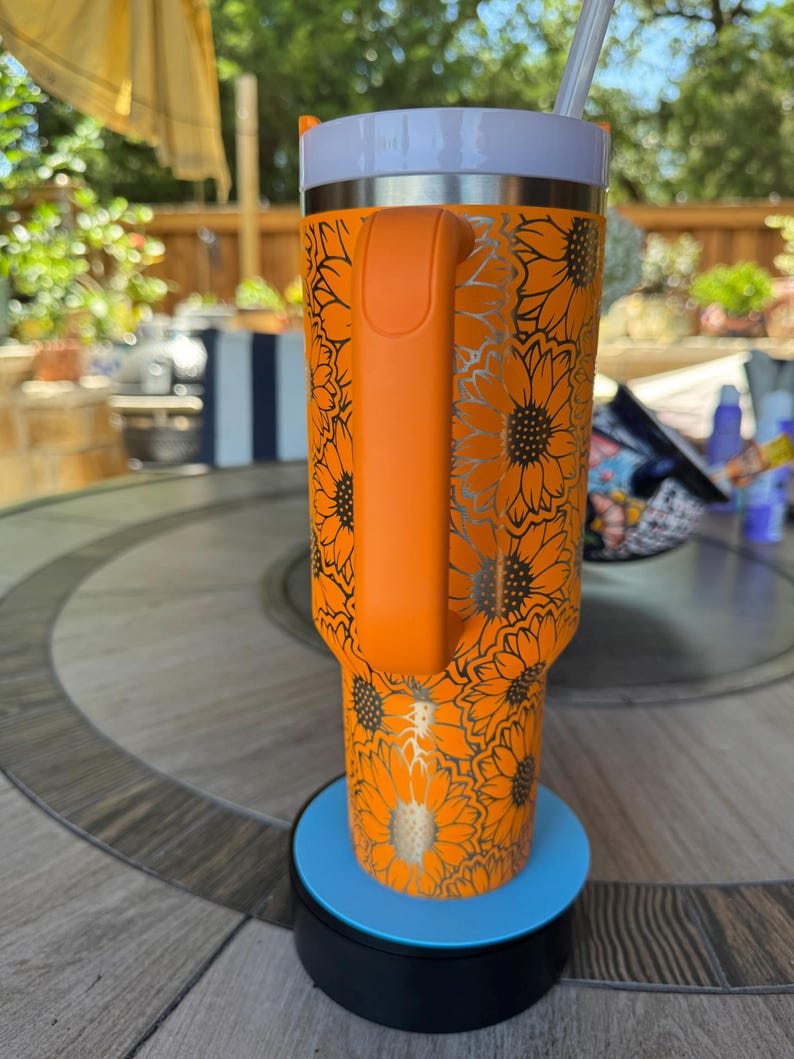 May include: A bright orange tumbler featuring a repeating sunflower pattern in a metallic silver finish. The cup includes a sturdy orange side handle, a white lid, and a clear straw. It sits upright on a circular blue and black base.