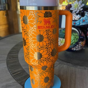 May include: A bright orange Stanley tumbler featuring a repeating sunflower pattern in a metallic finish. The cup includes a sturdy side handle, a white lid with an orange rotating cover, and a clear straw. The Stanley logo is printed in orange.