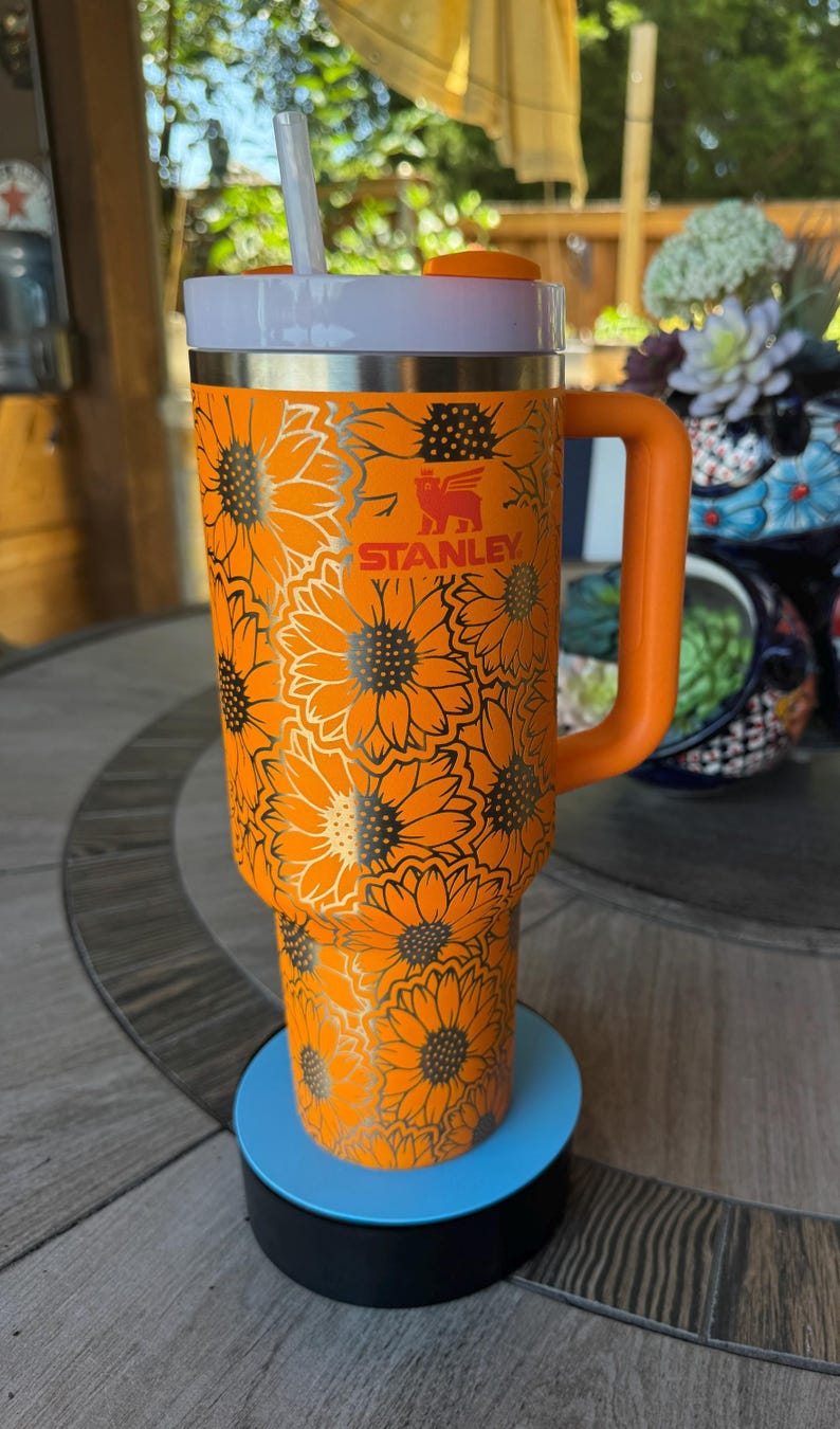 May include: A bright orange Stanley tumbler featuring a repeating sunflower pattern in a metallic finish. The cup includes a sturdy side handle, a white lid with an orange rotating cover, and a clear straw. The Stanley logo is printed in orange.