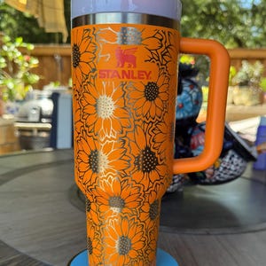 May include: A bright orange Stanley tumbler featuring a repeating black sunflower pattern across the exterior. The cup includes a matching orange side handle, a white lid with an orange rotating cover, and a clear straw protruding from the top centre.
