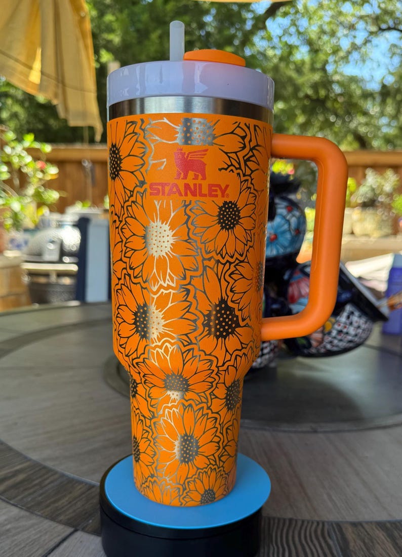 May include: A bright orange Stanley tumbler featuring a repeating black sunflower pattern across the exterior. The cup includes a matching orange side handle, a white lid with an orange rotating cover, and a clear straw protruding from the top centre.