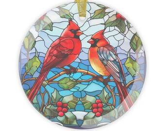 May include: Round ornament featuring a stained-glass design with two red cardinal birds perched on a branch. The background is a mosaic of blue and white, with green leaves and red berries. A gold ribbon is attached for hanging.